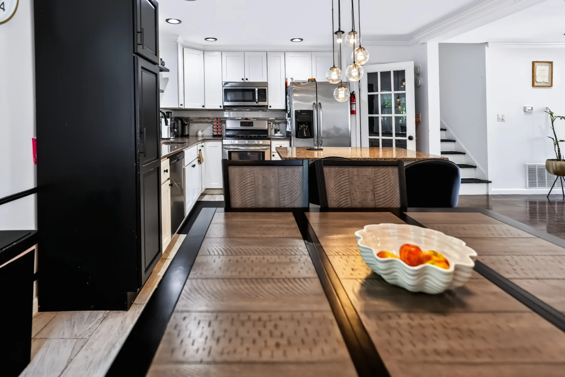 A bowl of fruit on a wooden dining table, with the kitchen beyond